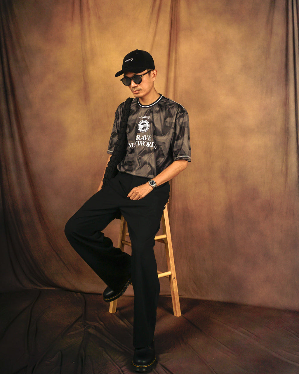 Model wearing a black SIMMY Brave New World oversized graphic jersey with a cap and sunglasses on a stool
